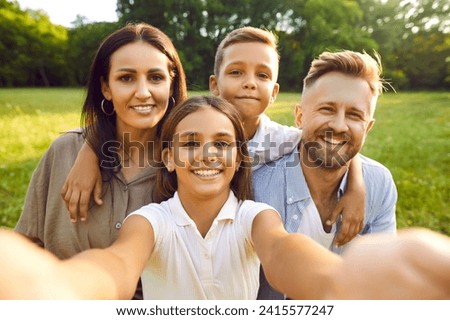 Happy family selfie in summer park. Parents and children having fun together. Pretty Caucasian european girl holds camera and takes photo of beautiful mother, handsome father and cute little brother