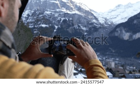 People vacationing in the Swiss Alps, man taking photo of two women with phone standing in chalet balcony during winter season with mountains in background covered in snow