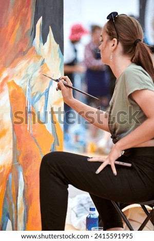Attractive female painter passionately draws picture with paintbrush sitting on chair for outdoor street exhibition, side view of female artist infusing life into outdoor art space