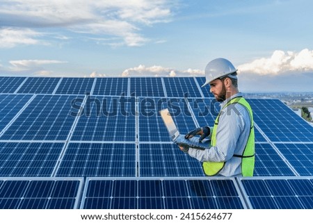professional engineer using a laptop in front of a solar panel power station, handsome male technician electrician looking at the screen holding a computer