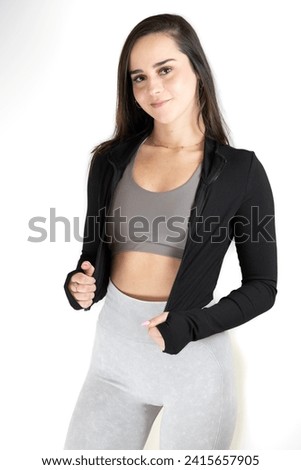 A young female adult stands in an active pose wearing a black yoga jacket, grey workout pants and gray sneakers, ready to start her fitness routine