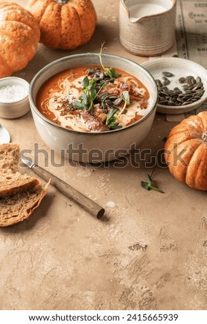 Bowl of pumpkin cream soup decorated with cream, pumpkin seeds, bacon, microgreens and olive oil on brown textured background with ingredients above. Vertical, text space