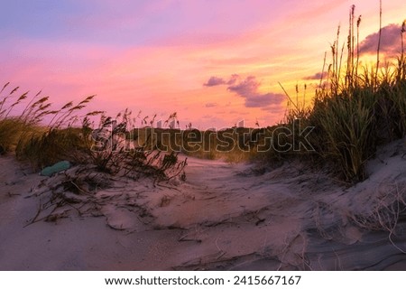 A tranquil sunset scene depicting an idyllic beach, with a sand dune and beach grass silhouetted against the soft, golden rays of the setting sun Royalty-Free Stock Photo #2415667167