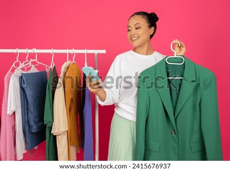 Beautiful asian woman holding green blazer on hanger and browsing app on her smartphone, happy korean female sharing or seeking fashion advice, standing against bright pink studio background