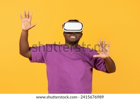 Virtual Reality Fun. Young black man engrossed in online simulation game via VR headset, raising hands playing immersive videogames over yellow studio background, portrait shot