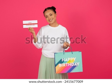 Beautiful asian woman holding gift certificate and 'Happy Birthday' shopping bag, happy young korean lady enjoying bday offer and loyalty program, standing on pink studio background, copy space