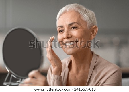 Anti Aging Skincare. Cheerful Senior Lady Applying Facial Moisturizer Preventing Wrinkles Looking At Round Mirror, Caring For Her Ageless Beauty At Home, Enjoying Self Pampering Royalty-Free Stock Photo #2415677013