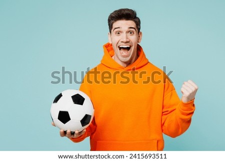 Young happy man fan he wears orange hoody casual clothes do winner gesture cheer up support football sport team hold in hand soccer ball watch tv live stream isolated on plain blue color background Royalty-Free Stock Photo #2415693511