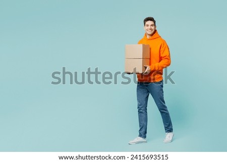 Full body smiling happy fun young man he wears orange hoody casual clothes hold stack cardboard blank boxes isolated on plain pastel light blue cyan color background studio portrait. Lifestyle concept