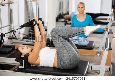 Young and elderly women wearing activewear and doing reformer exercises on Pilates machine in rehabilitation center