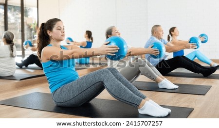Sporty young girl doing sit-ups with bender ball to strengthen abs muscles during group pilates class in fitness studio Royalty-Free Stock Photo #2415707257