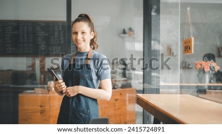 Beautiful young barista woman in apron holding tablet and standing in front of the door of cafe with open sign board. Business owner startup, SME entrepreneur seller business concept.