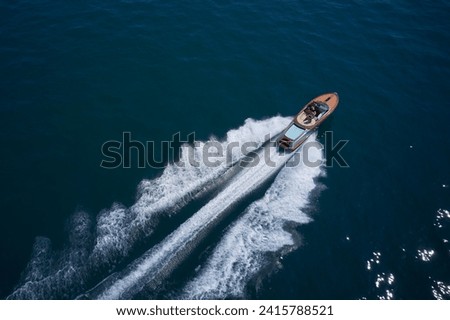 Expensive wooden boat, man and woman in motion on the water making a white trail looking like air. A large modern high-speed wooden luxury boat moves on blue water, top view. Royalty-Free Stock Photo #2415788521