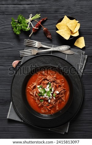 birria de res, mexican beef stew in hot red pepper sauce with raw onion and chopped cilantro, taco chips in black bowl on black wooden table with forks, vertical view