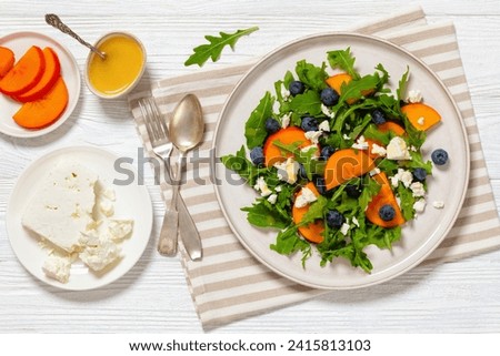 arugula persimmon white greek cheese and blueberry salad with honey and olive oil dressing on plate on white wooden table with ingredients, horizontal view from above, flat lay