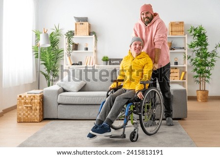 Portrait of a caregiver and a disabled man in wheelchair looking and smiling at camera at home Royalty-Free Stock Photo #2415813191