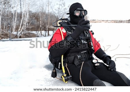Portrait of a man wearing special wetsuit, mask, gloves, mask and flippers, sitting on the snowy edge of an ice hole, ready to immerse underwater in winter, equipped and tightened with a safety rope Royalty-Free Stock Photo #2415822447