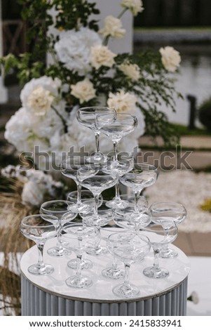 Pyramid of champagne glasses at a party. Wedding decorations in luxury ceremony. Location for celebration on pier. White arch for ceremony is decorated flowers and greenery in backyard with lake view.