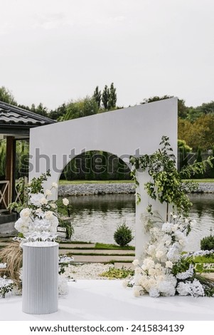 Location for celebration on pier. Wedding decor in banquet area. White arch for ceremony is decorated flowers and greens, greenery in backyard with lake view. Pyramid of champagne glasses at a party.