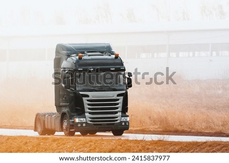 An empty truck with a modern cabin drives on a speedway. The truck has no trailer connected and is carrying no load. The road is bordered by wild grass and a long horizontal structure.