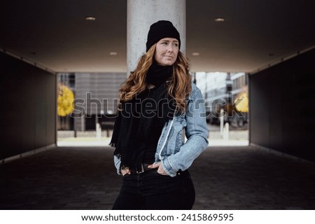 The picture shows a woman around thirty with long wavy red hair. She is dressed with black cap denim jacket and black pants scarf and sweater.