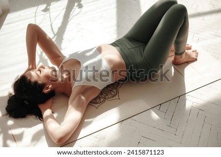 Caucasian dark-haired lady lying on white fitness mat under sun rays indoors. Carefree female in comfortable sports clothing practicing yoga with closed eyes and relaxing. Royalty-Free Stock Photo #2415871123