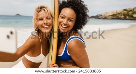 Two young women in bikinis, one holding a surfboard, take a beach selfie with a camera phone. They happily celebrate their friendship and create lasting memories on a fun surfing trip.