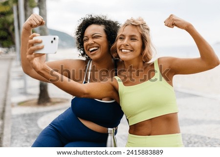 Two women in sportswear take a selfie with a smartphone, smiling and showing off their healthy biceps. Friends celebrating a healthy lifestyle after a great workout on the ocean promenade.