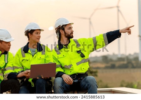 Close up technician worker man point to right side and stay with his co-worker using laptop to discuss work in front of windmill or wind turbine with evening light.