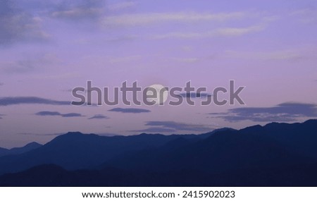 white full moon rising behind a mountain ridge illuminating misty clouds creating a mystical atmosphere. glowing round moon rising into the night sky with layers of hills. luna above peak. night scene
