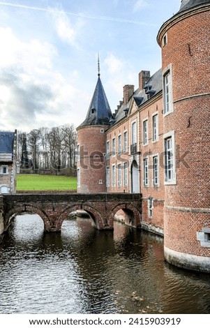 Arched bridge over moat surrounding 16th century Alden Biesen castle, circular towers and gable roof, bare trees in background, cloudy day in Bilzen, Limburg, Belgium