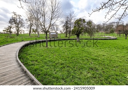 Flanders organic orchard at Alden Biesen Castle with curved wooden bridge over green grass, bare fruit trees against overcast sky in background, cloudy winter day in Bilzen, Limburg, Belgium Royalty-Free Stock Photo #2415905009