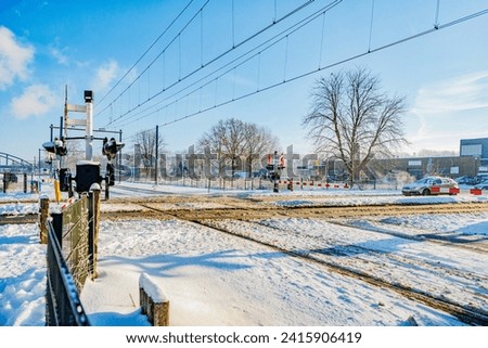 Snow covered railway crossing on road in winter landscape, closed downward barriers, stop sign and warning lights, stopped car waiting in background, sunny day in Beek, South Limburg, Netherlands