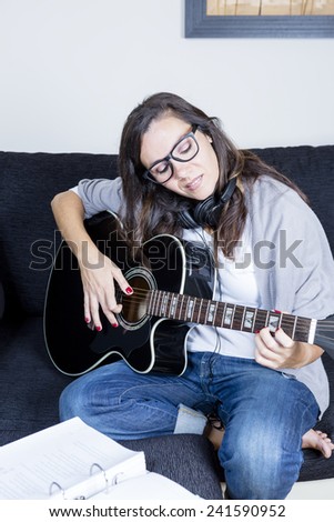 smiling young woman is playing a black acoustic guitar sitting on a sofa at home with headphones and sheet music book - focus on the girl 
