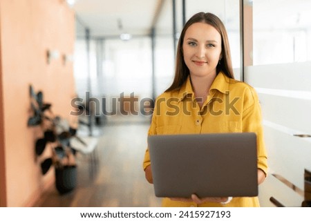 Smiling woman with laptop looks at camera and standing indoor in modern office, cheerful businesswoman in casual wear checking emails standing in hallway