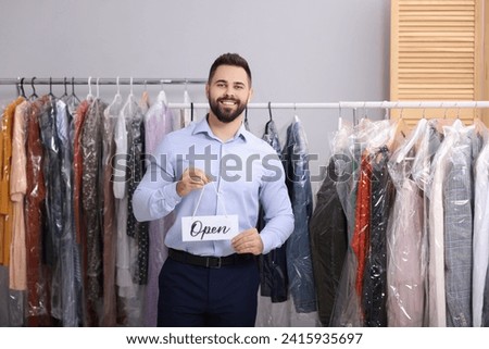 Dry-cleaning service. Happy worker holding Open sign near racks with clothes indoors