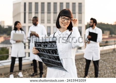 Multiethnic employees of clinic working on devices with results of examination of patients. Portrait of Caucasian doctor holding x-ray scan of patient during break outdoors hospital looking at camera.