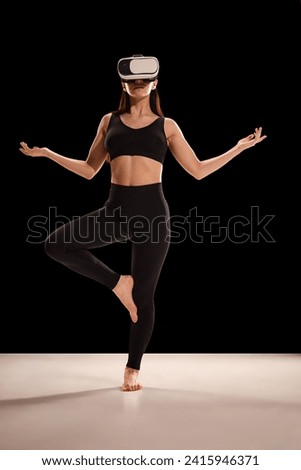 Woman in fitness costume and VR glasses doing yoga exercises against black studio background. Body care and mindfulness. Concept of sport, dieting, body care and active lifestyle.
