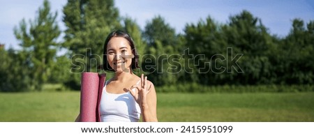 Excited young woman standing with sports mat, yoga clothes, shows okay sign, workout in park, wellbeing training session outdoors.
