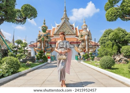 Young woman wearing traditional Thai dress stands holding an antique bag with giant front ornaments at the Wat Arun Temple, a popular destination for tourists around the world. Bangkok Thailand