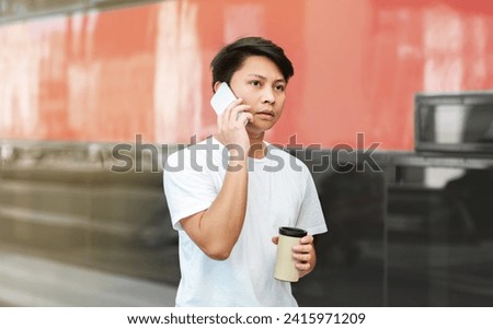 Concentrated asian guy tourist talking on phone while waiting for train at station, drinking takeaway coffee, looking at copy space for advertisement. Tourism, vacation, journey, weekend trip