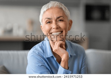 Portrait of lovely older gray woman smiling to camera and touching chin, sitting on sofa at home indoors. Happy beautiful mature lady resting and relaxing in living room, expressing happiness Royalty-Free Stock Photo #2415971253