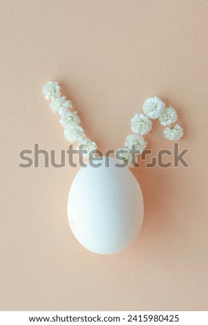 vertical photo of a top view of a white egg with ears like the Easter bunny made from white flowers on peach fuzz background. Happy Easter concept. soft focus