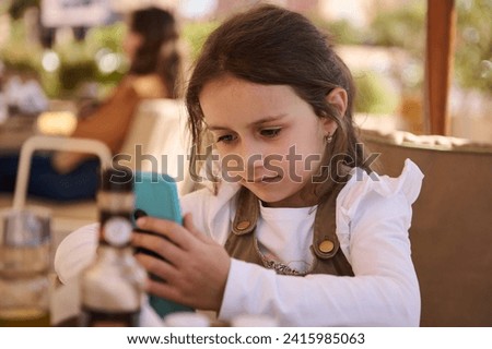 Adorable Caucasian little child girl sitting at table, having fun on mobile phone, playing online video games, watching cartoons. Digital gadget addiction. People. Children. Technologies