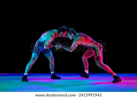 Young athlete man, wrestlers in blue and red uniform hand wrestling in neutral position on their feet in neon lights against black background. Concept of fair wrestling, championship, win competition.