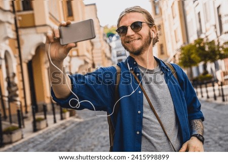 Handsome young man in casual clothing smiling while taking selfie outdoors. Caucasian tourist talking with friends by smartphone while walking on city streets. Vlogging