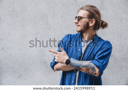 Smiling young nerdy bearded stylish man hipster freelancer is standing on pure background in sunglasses and casual outfit, pointing on the copyspace
