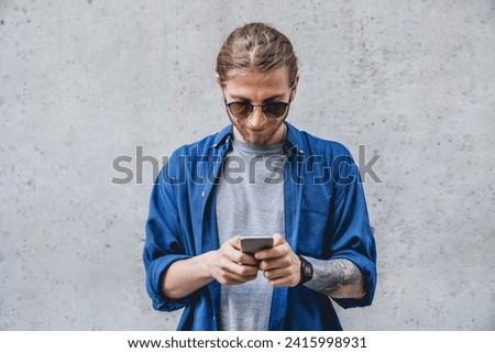 Young man texting message on smart phone isolated on grey background. Smiling caucasian male in sunglasses holding smartphone and looking at it.