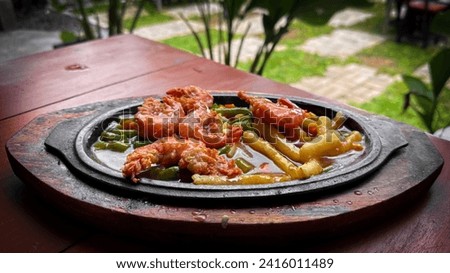 landscape picture of  Crispy Shrimp Steak on the restaurant with blurry plant background. there's French fries and vegetables. The steak is served with sauce on a hot plate on a wooden board