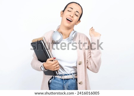 Overjoyed successful beautiful arab student holding notebooks raises palm and closes eyes in joy being entertained by friends